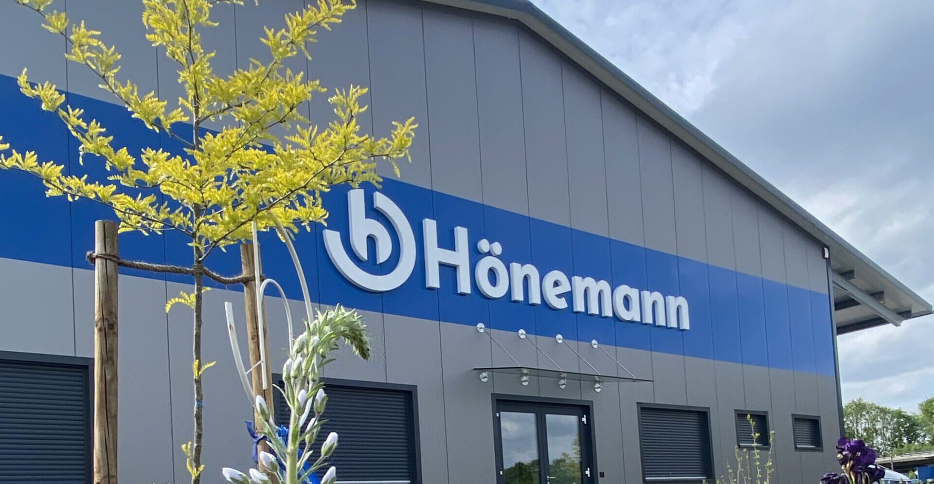 Das Foto zeigt das Firmengebäude der Hönemann GmbH, ein moderner Unternehmenssitz mit großzügiger Glasfront und klaren architektonischen Linien. Das Gebäude befindet sich auf einem gepflegten Firmengelände und trägt gut sichtbar das Firmenlogo am Haupteingang. Es repräsentiert die professionelle Ausrichtung und das zukunftsorientierte Selbstverständnis des Unternehmens. Firmengebäude der Hönemann GmbH mit moderner Fassade und Firmenlogo am Eingangsbereich.