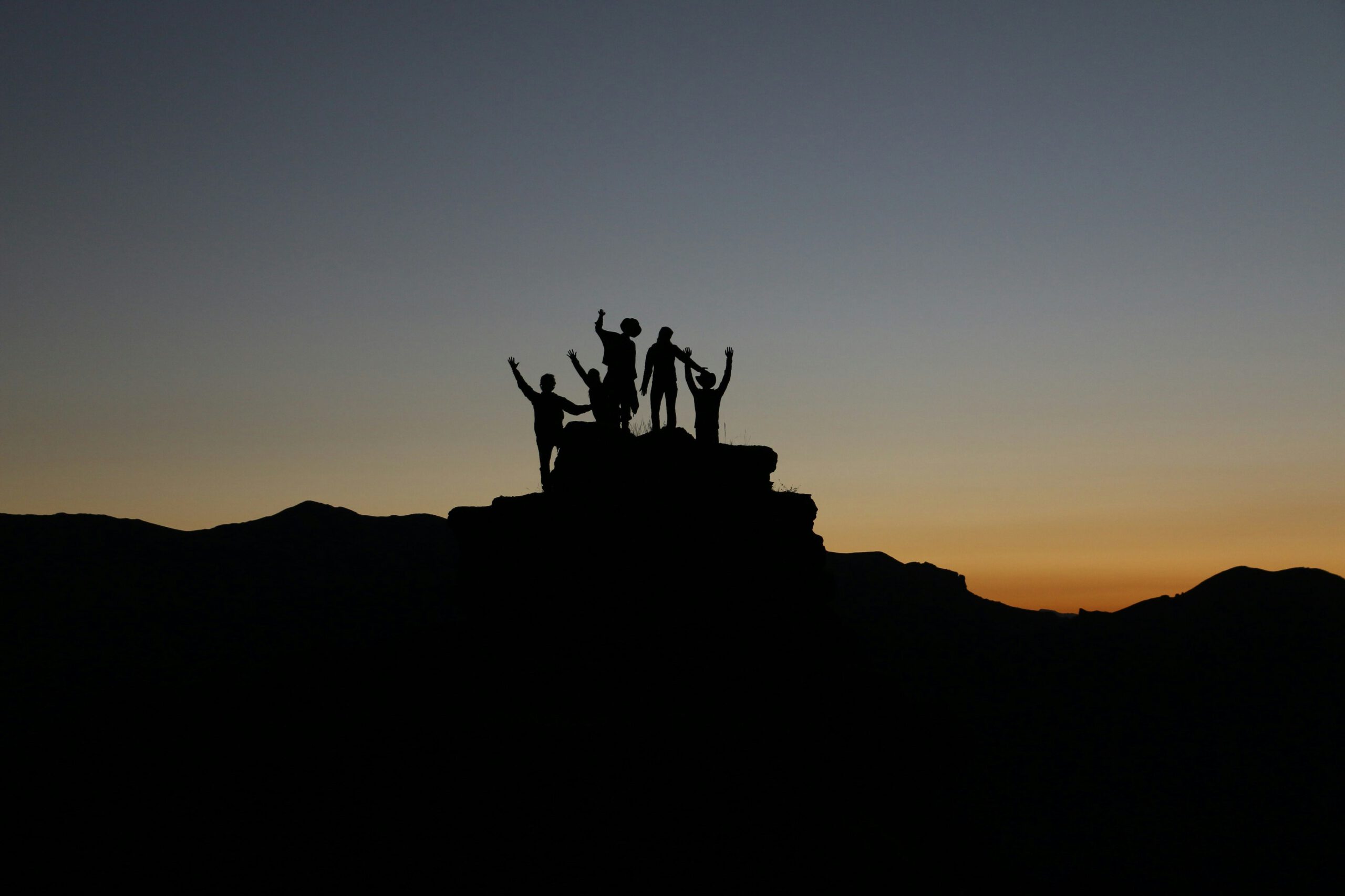 Silhouetten von Menschen auf einem Berggipfel im warmen Licht der Abendsonne – Symbol für Gemeinschaft, Zusammenhalt und soziale Verantwortung.